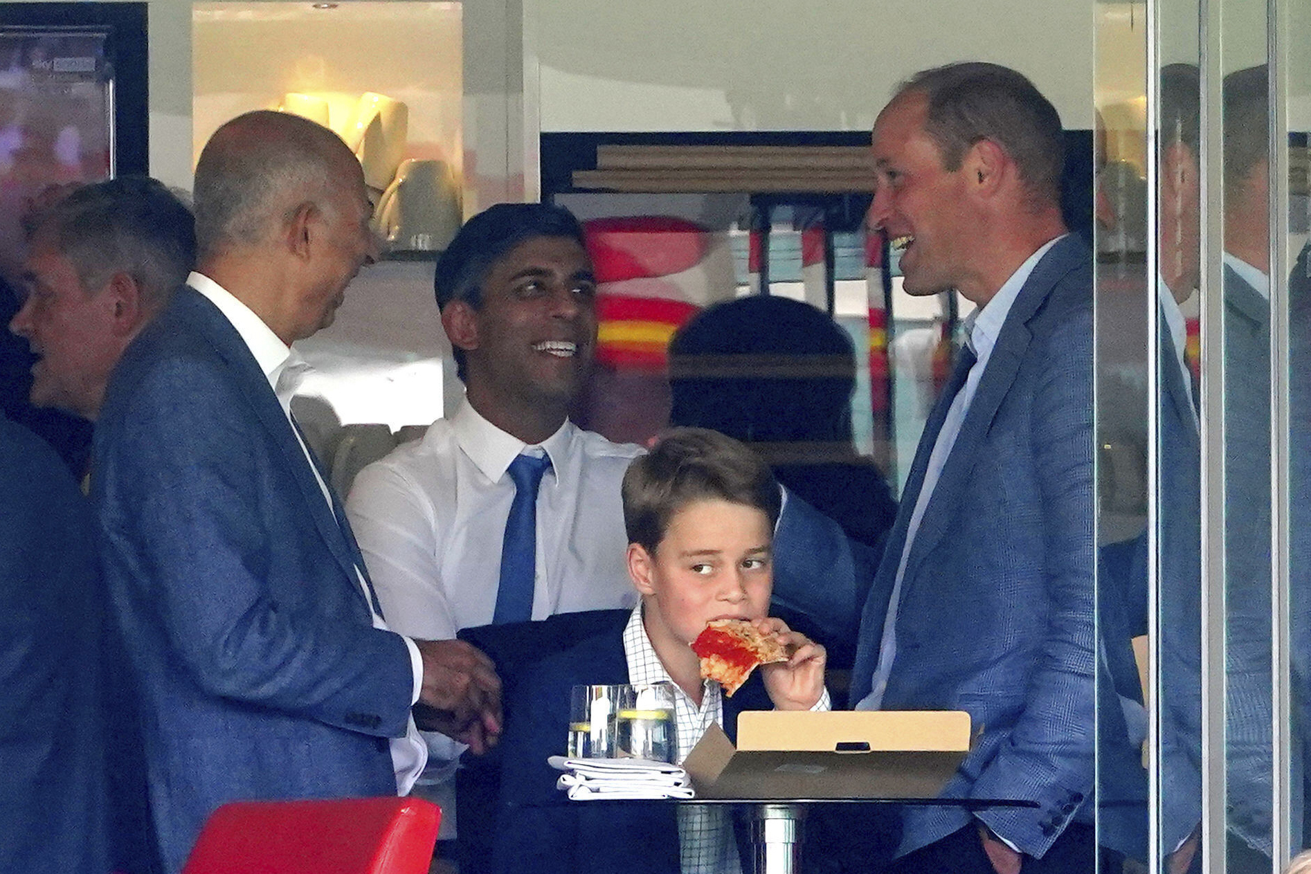 Three men in formal wear speak to each other as a young boy eats pizza in the foreground.