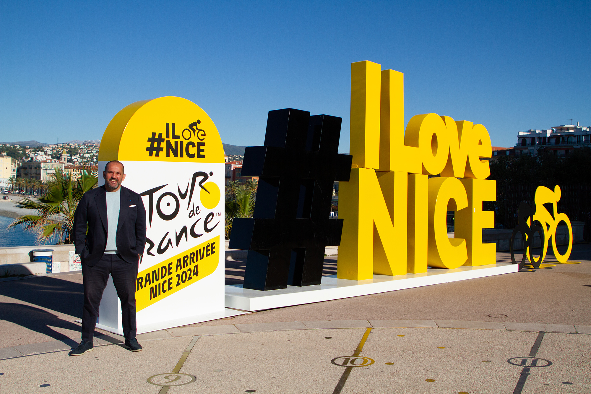 A man stands in front of a bright yellow 'I Live Nice' sign with Tour de France signage on it.