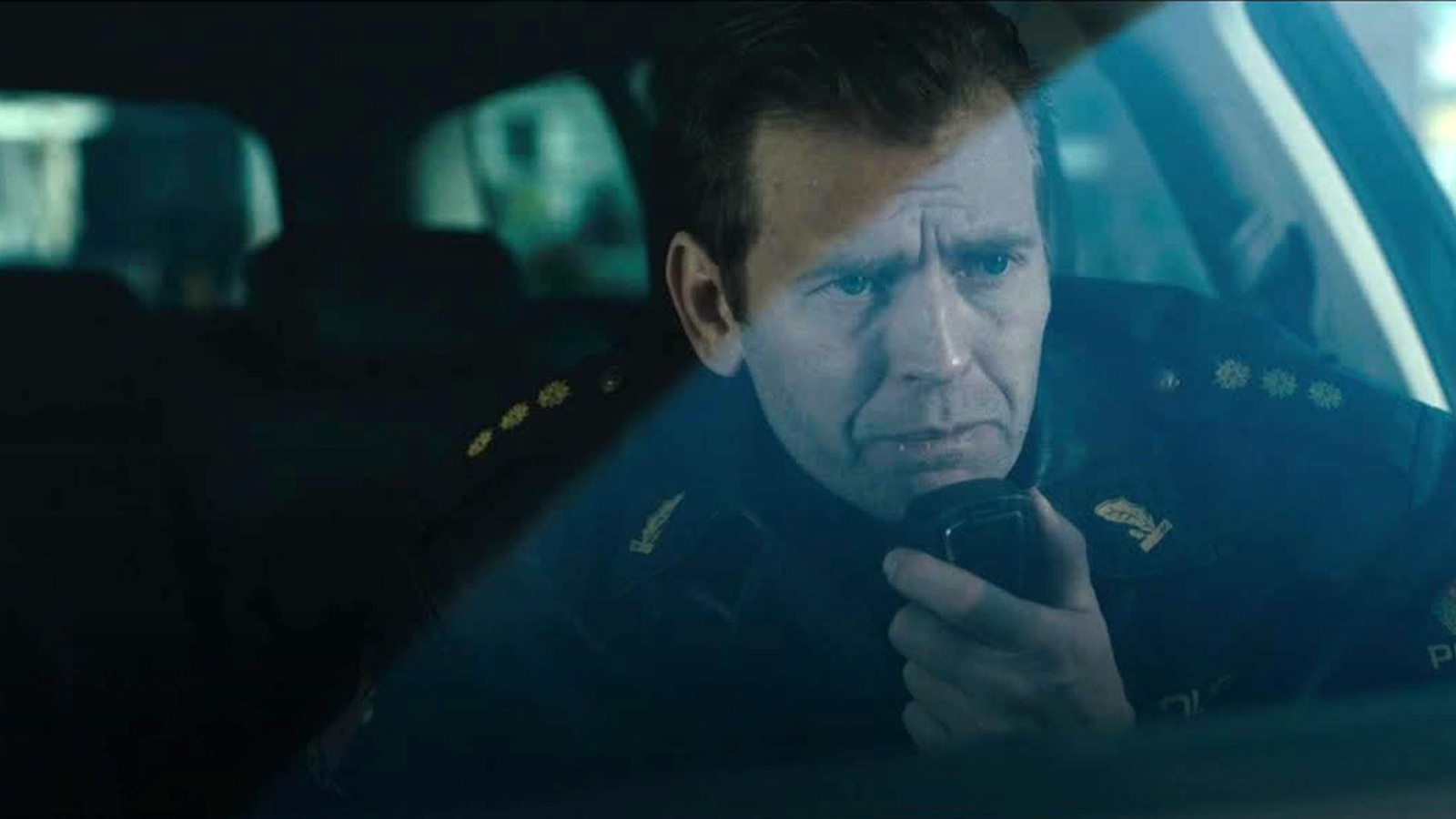 A policeman sits in a car, speaking into a hand-held. 