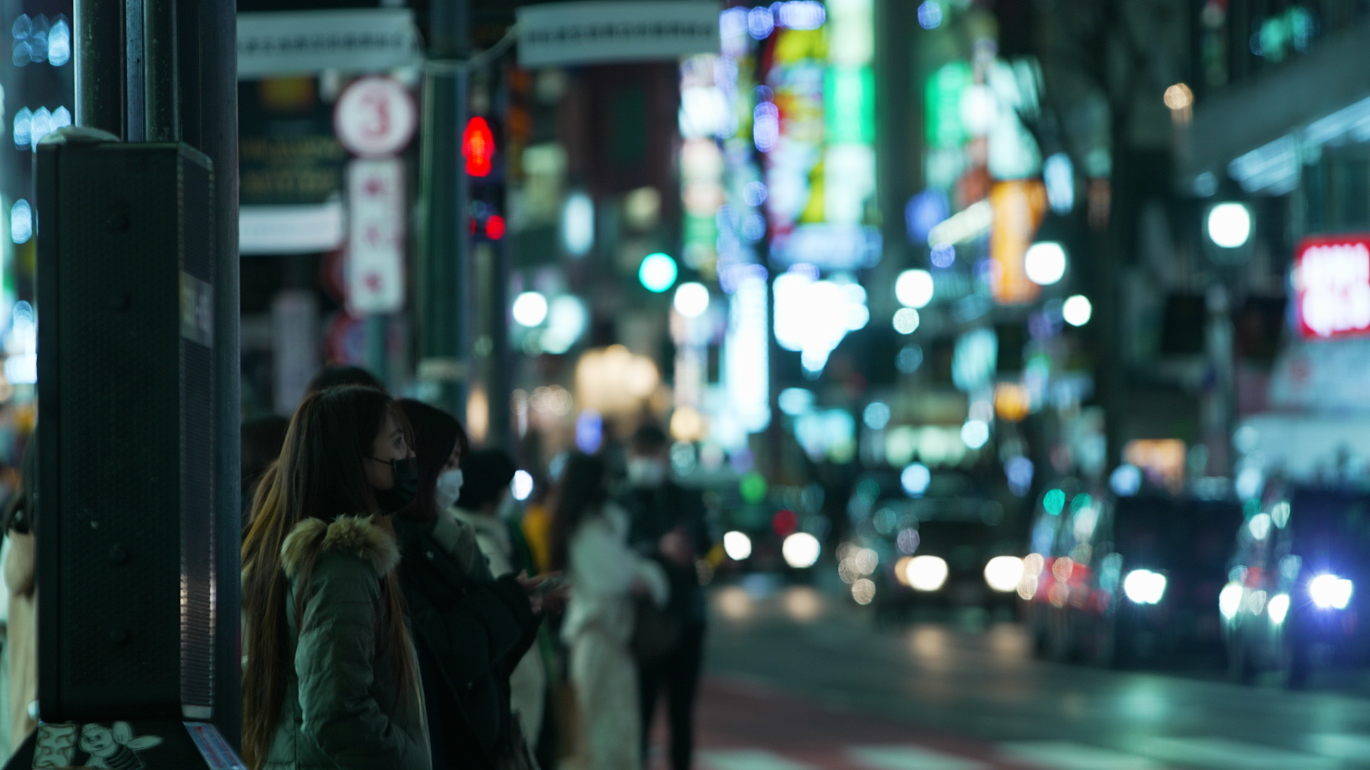 People stand on the side of a road in Japan with lots of bright lights in the background.