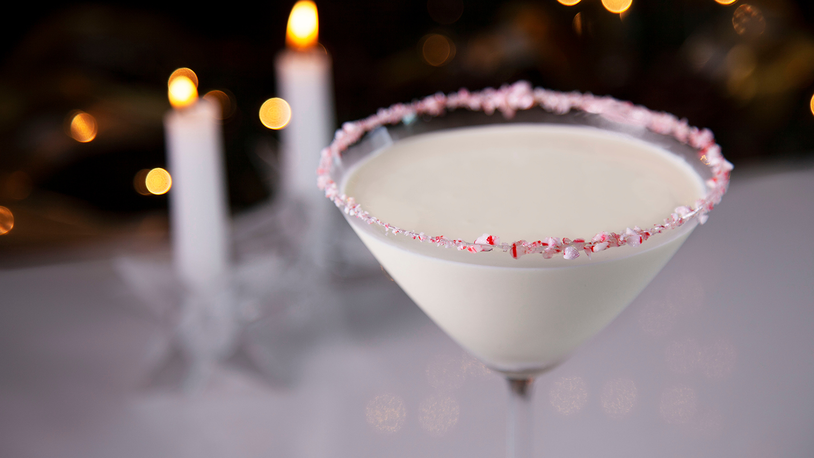 A martini glass rimmed with crushed candy cane holds a white cocktail. Two lit candles can be seen behind the glass, out of focus. 