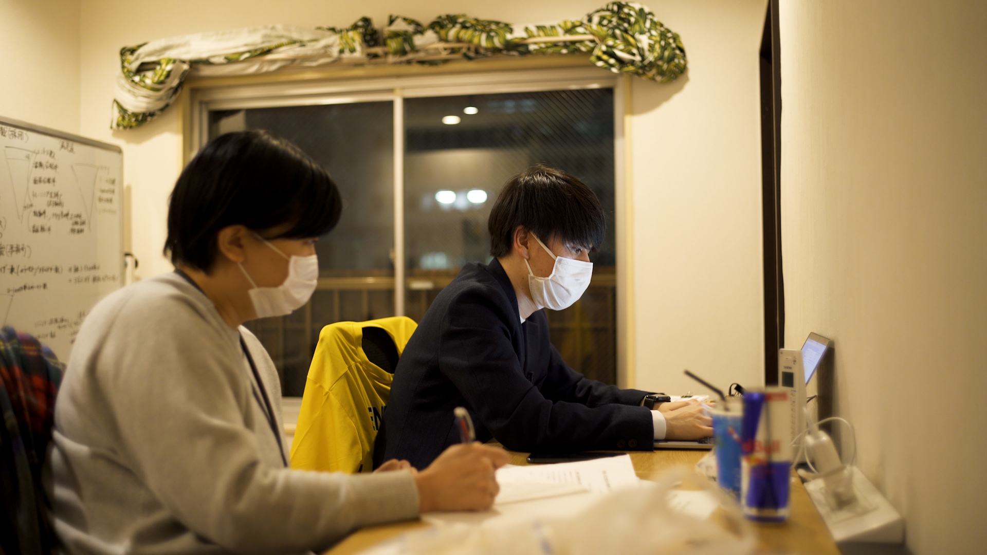 Two people sit at computers wearing masks.