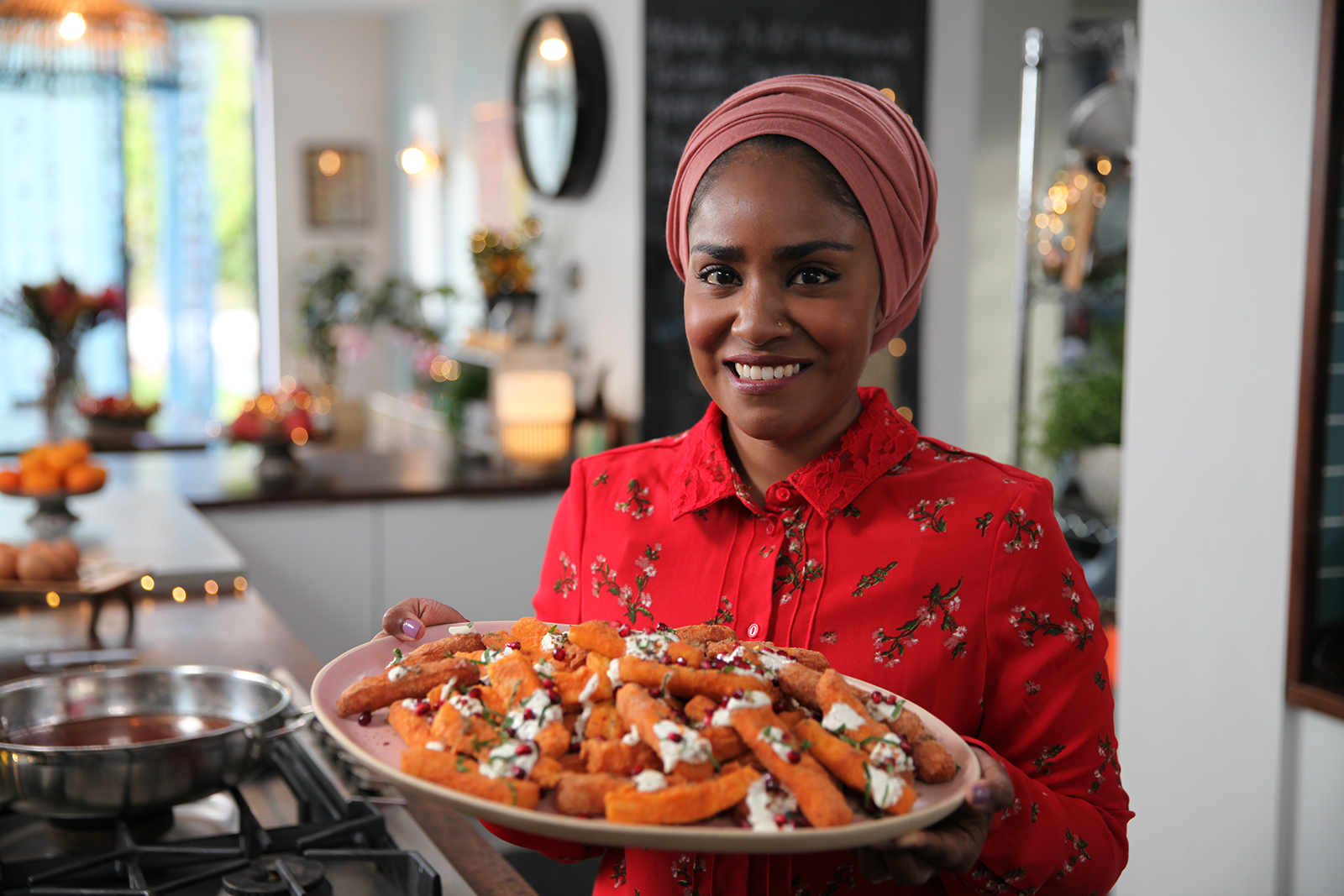 A woman in a bright red top and pink headscarf stands in a kitchen, smiling, holding a platter.