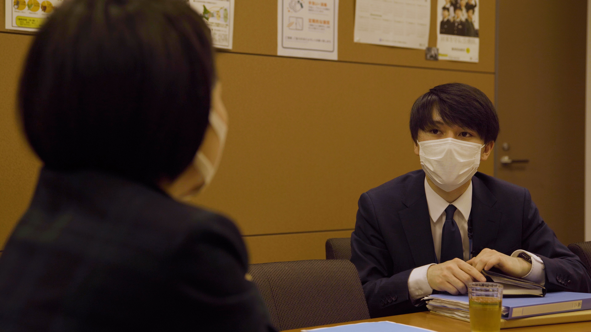 A young man wearing a mask looks at another person in the foreground.