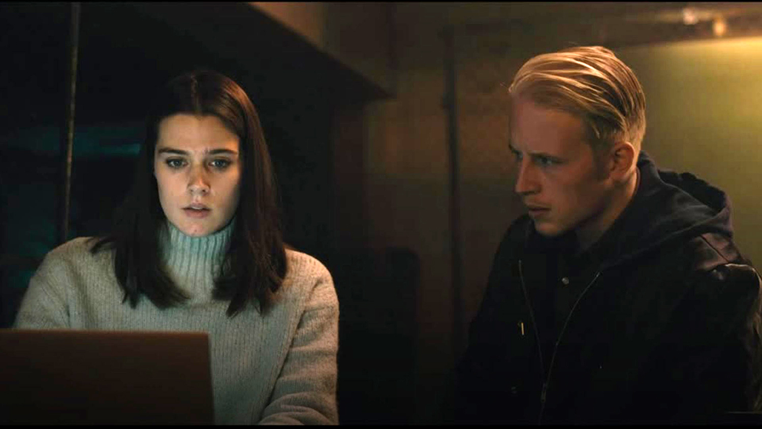 A young woman sits at a laptop in a dark room, with young man setting beside her, looking at her with a worried expression.