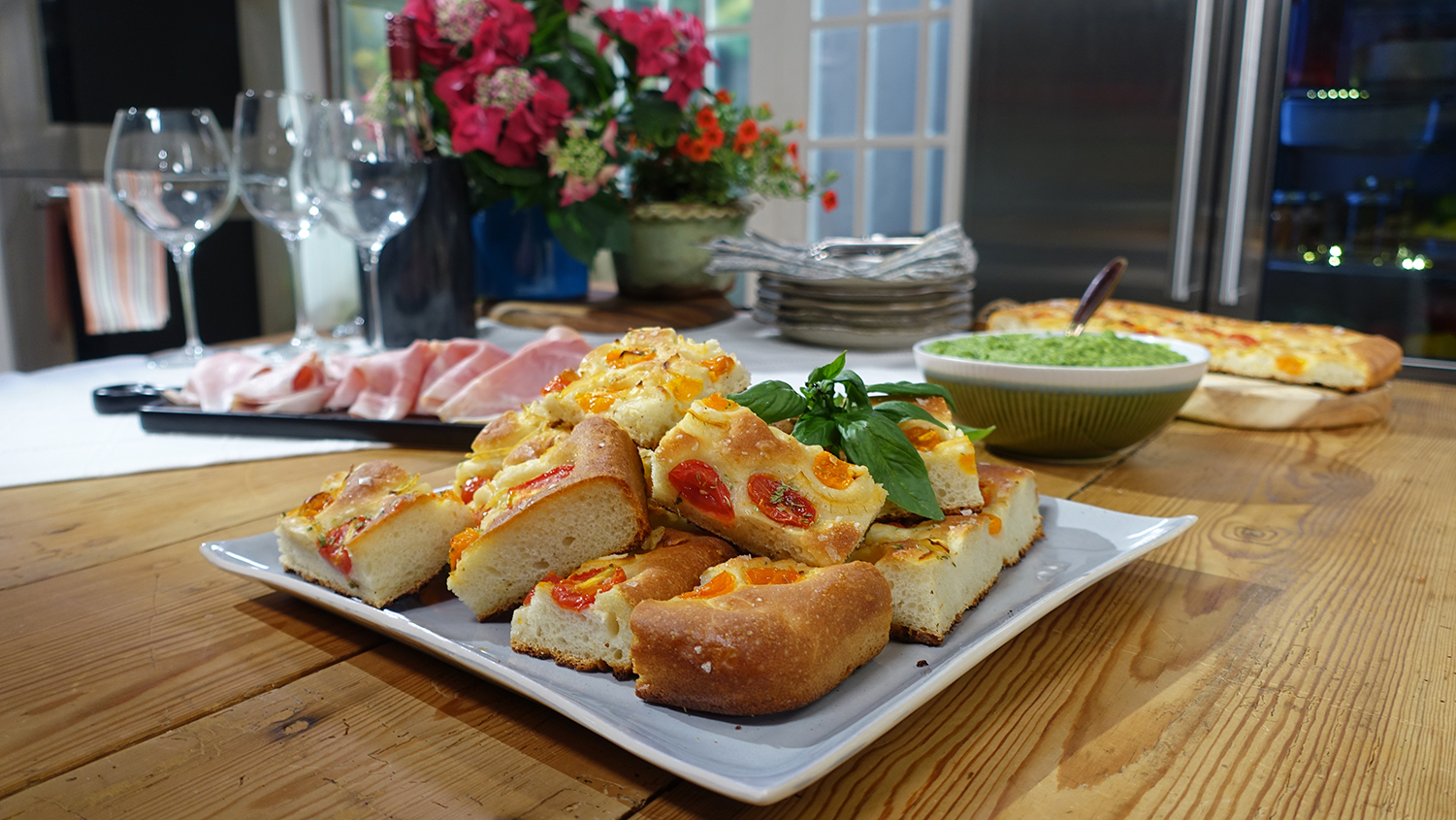 A platter with chunky pieces of focaccia, studded with orange and red tomato, sits on a wooden table. More food, glasses and plates can be seen behind the platter. 