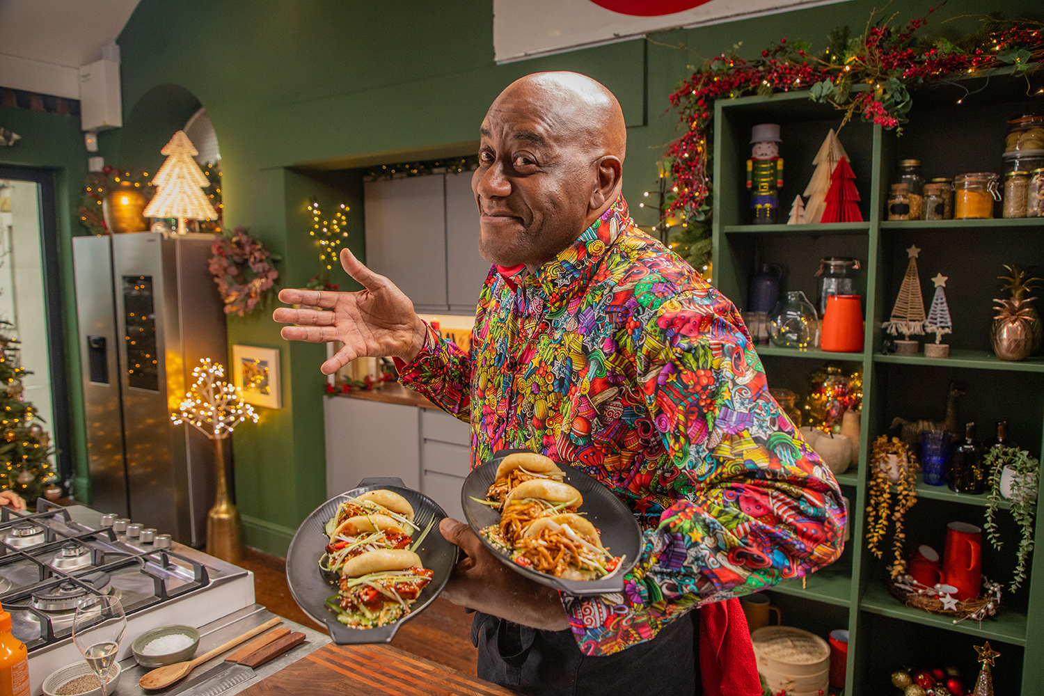 Ainsley Harriott in a very bight shirt stands in a kitchen, holding two plates with filled split buns. He's smiling and gesturing with his free hand. 