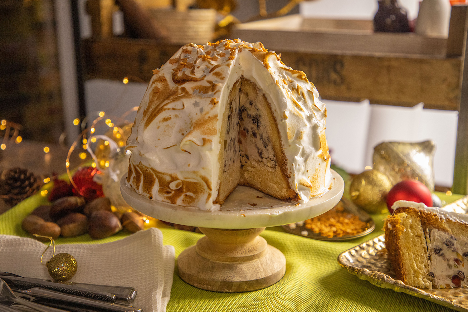 A dome coated in cooked meringue with a slice out to show the interior, sits in a footed stand amid festive table decorations. 